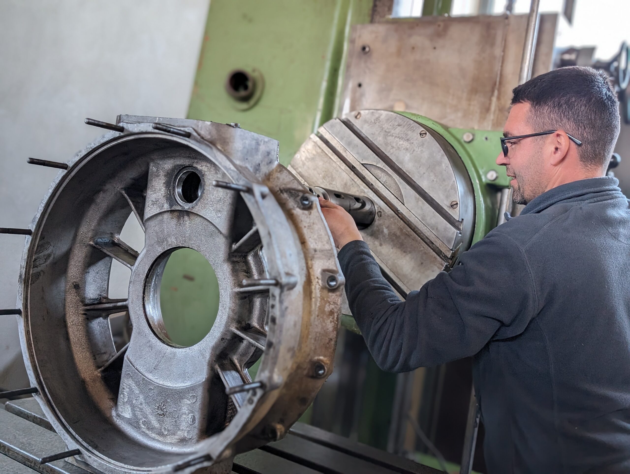 Na slici se vidi majstor Đokić Dragiša koji radi na CNC mašini za obradu metala. Majstor pažljivo obrađuje veliki metalni deo, fokusirajući se na preciznost i detalje. Mašina i okruženje ukazuju na visokotehnološku radnu sredinu specijalizovanu za kompleksne zadatke obrade metala.